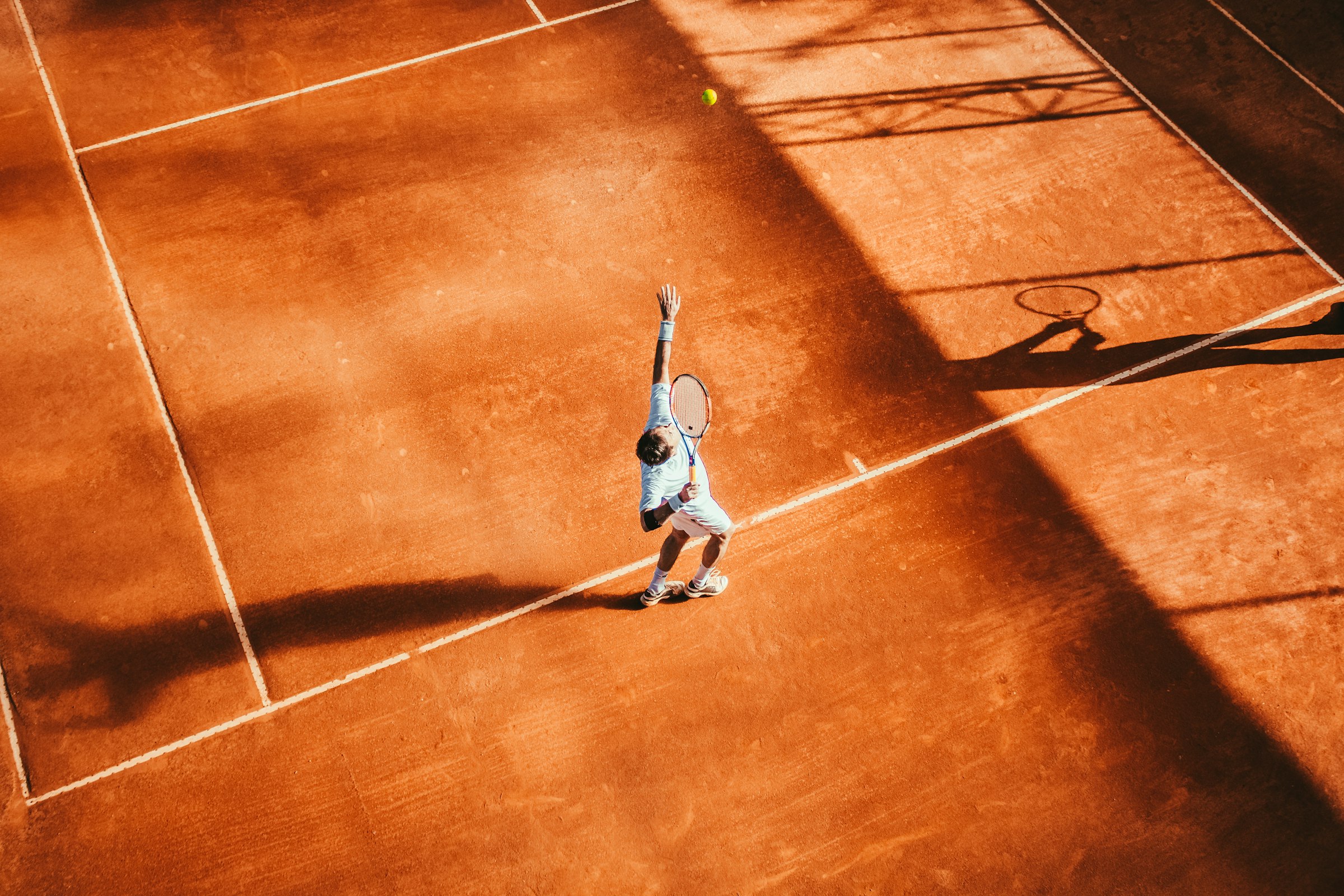 A player serving on a clay court, seen from above in late-afternoon light